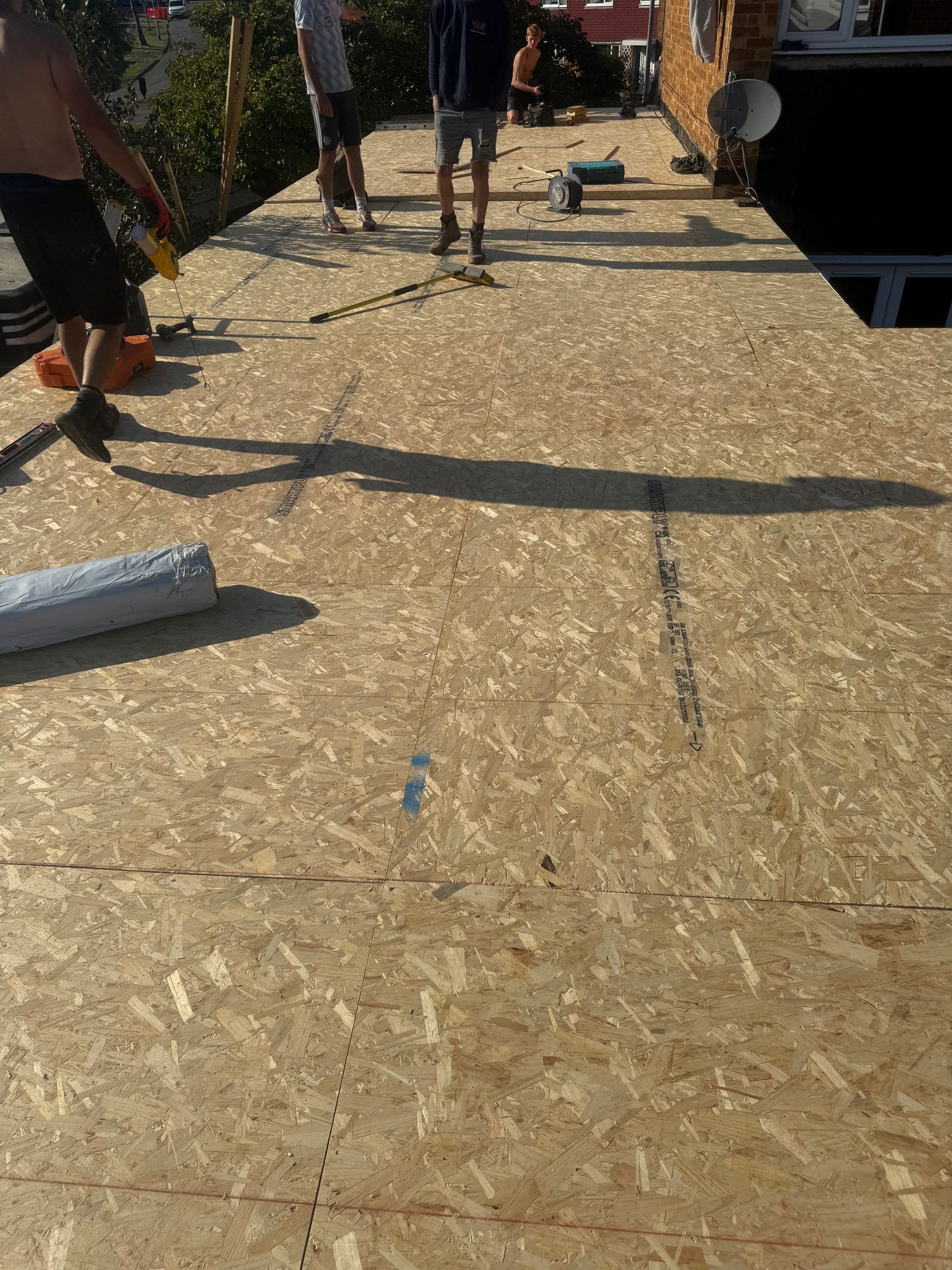 Workers installing OSB roof decking across a flat roof area, with tools and construction materials visible on the surface