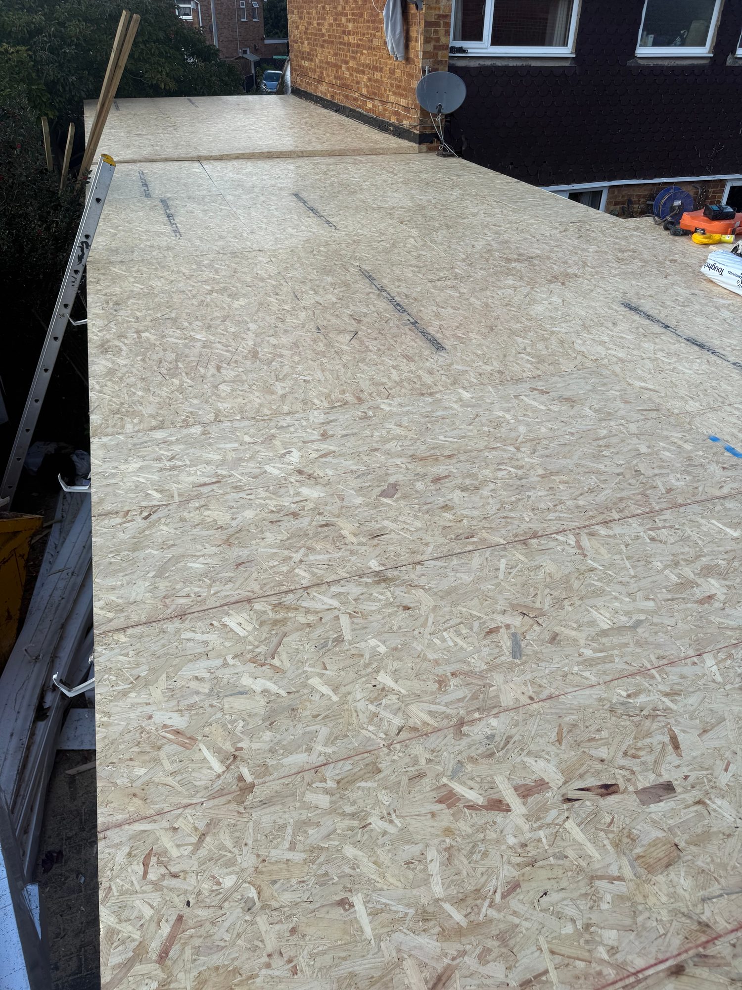 OSB decking panels covering the roof area beside the brick building, with a ladder and construction tools visible at the edges