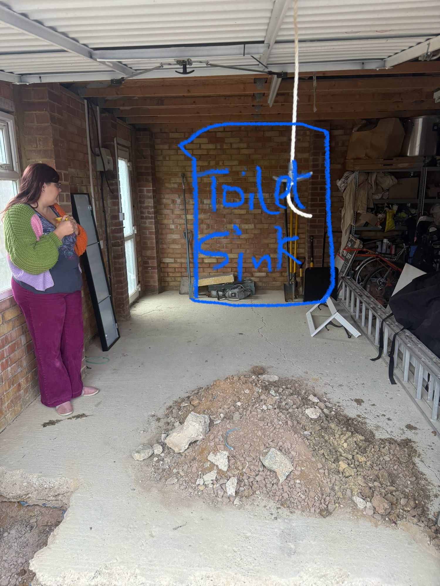 Interior of a brick garage with exposed timber roof framing, windows and stored tools, with a blue outline marking an area labelled for a toilet and sink and a pile of broken concrete and rubble on th