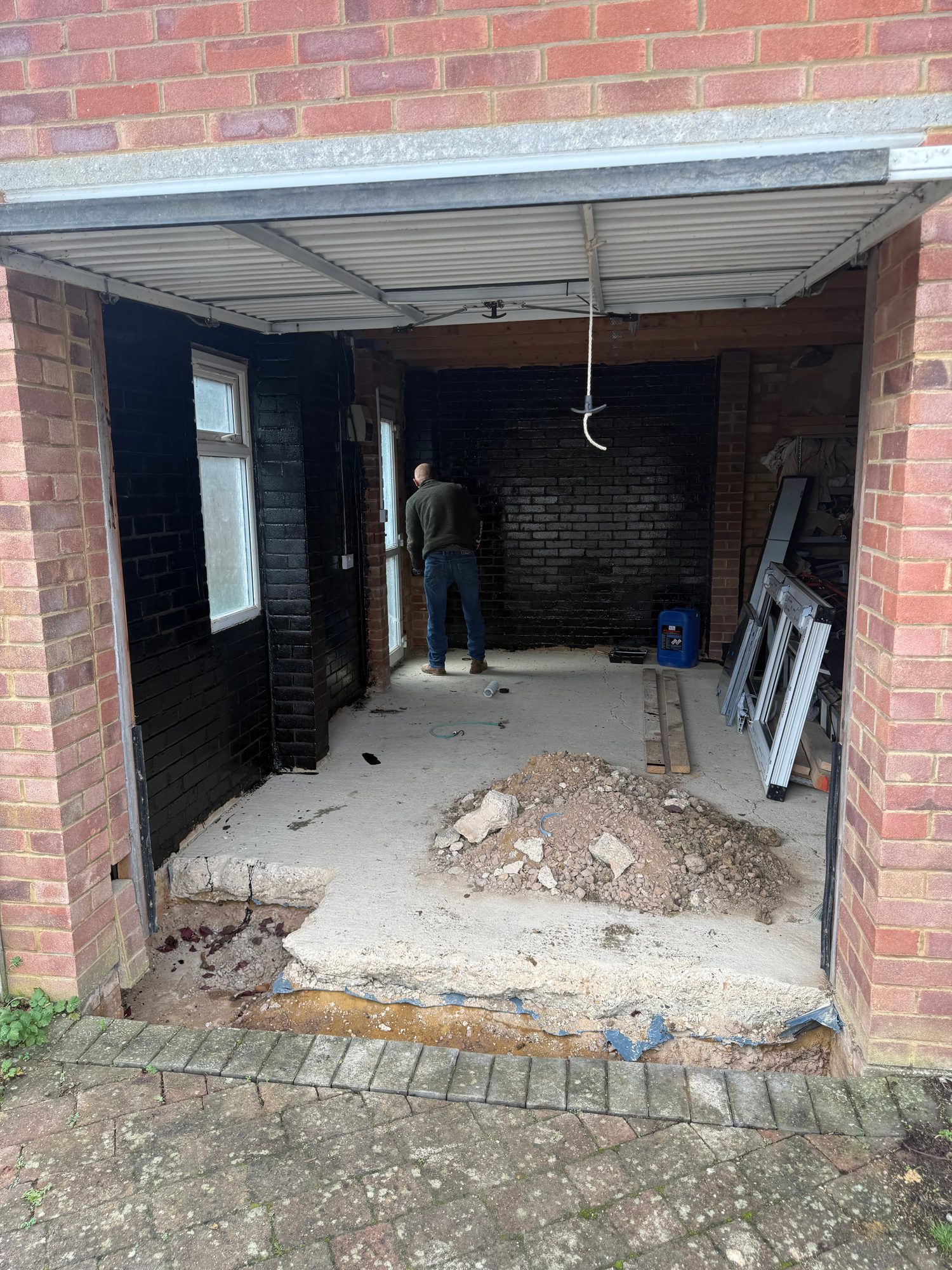 Garage interior before renovation, showing exposed brick walls, a concrete floor with a pile of rubble, stored tools and materials, and a partially open garage door