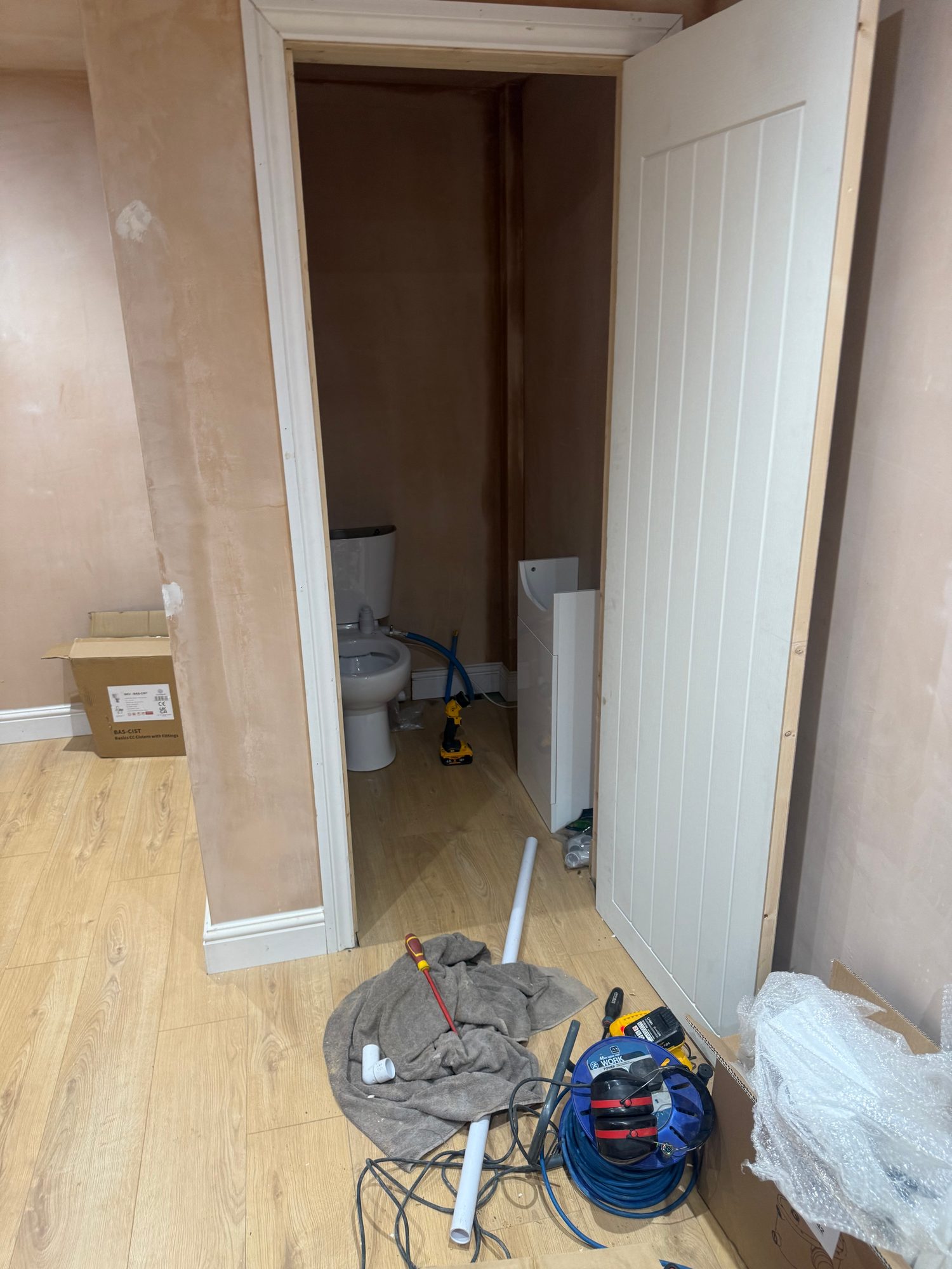 Bathroom with wood-effect flooring installed, an open white door, visible toilet and plumbing components, with tools and materials on the floor during ongoing works