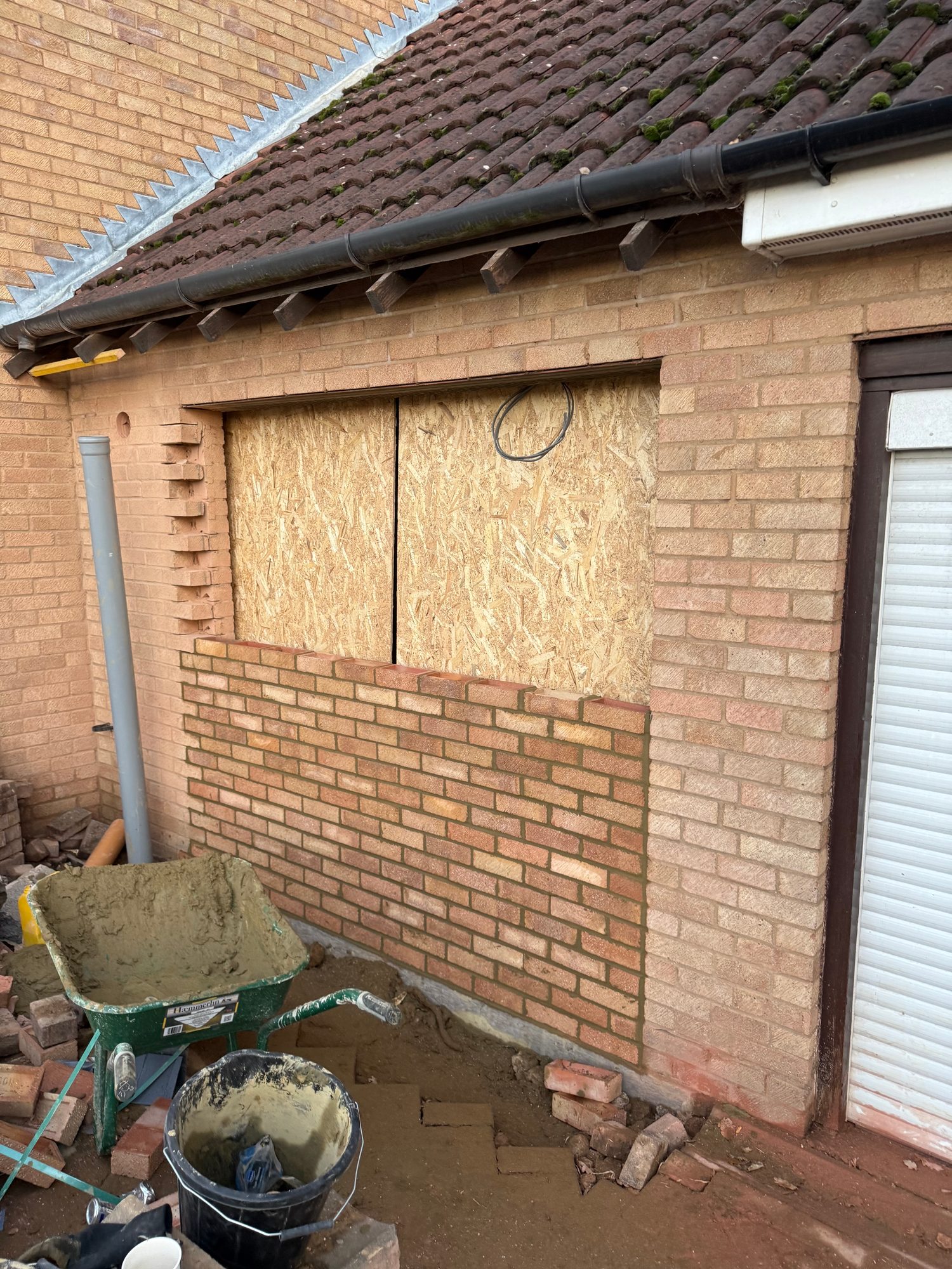 Brick garage wall with a boarded-up opening, partially completed brickwork below the opening, roof tiles and guttering above, and building materials and tools on the ground