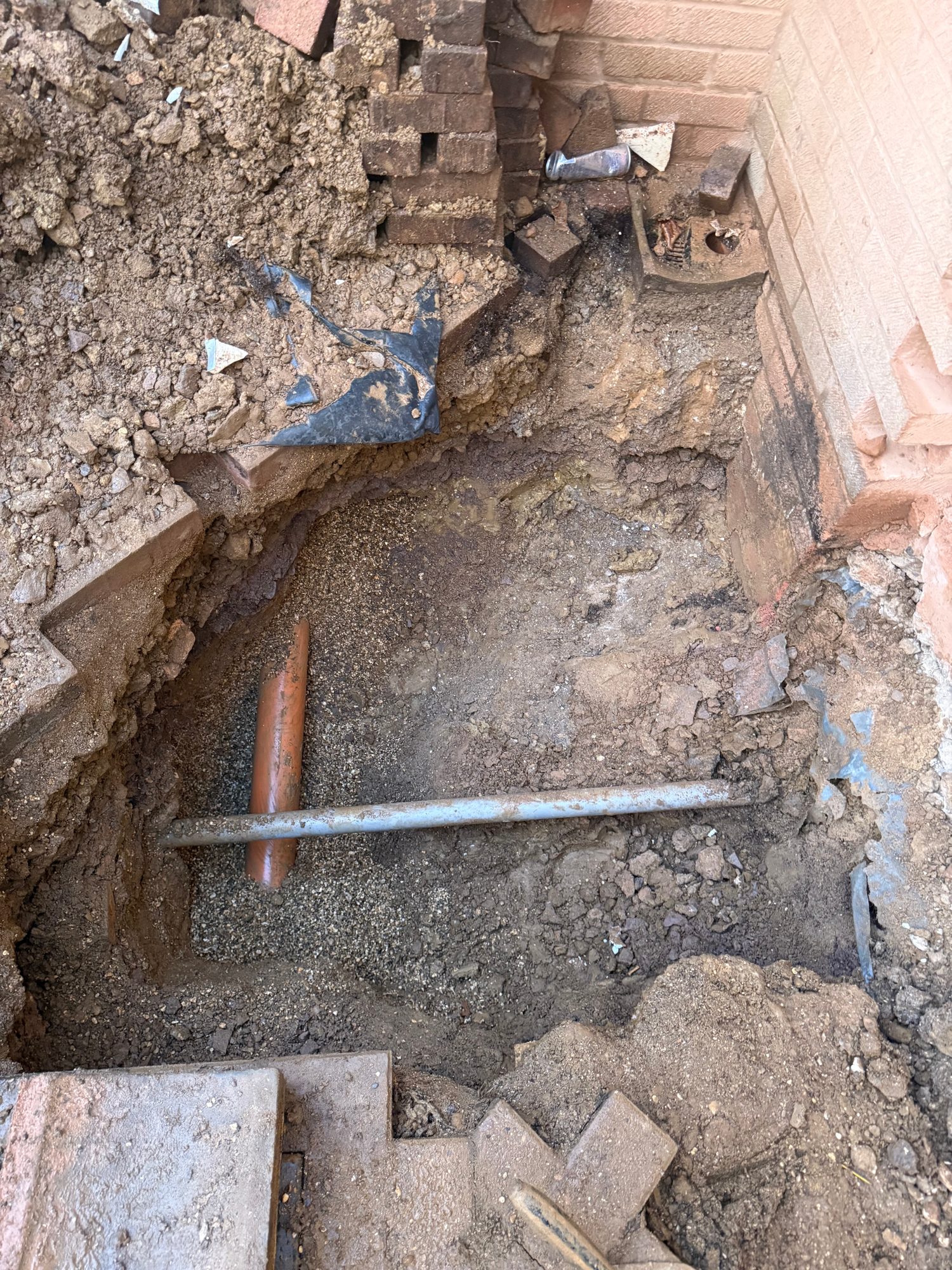 Excavated area beside a brick wall showing exposed soil, loose bricks and two visible pipes within the ground