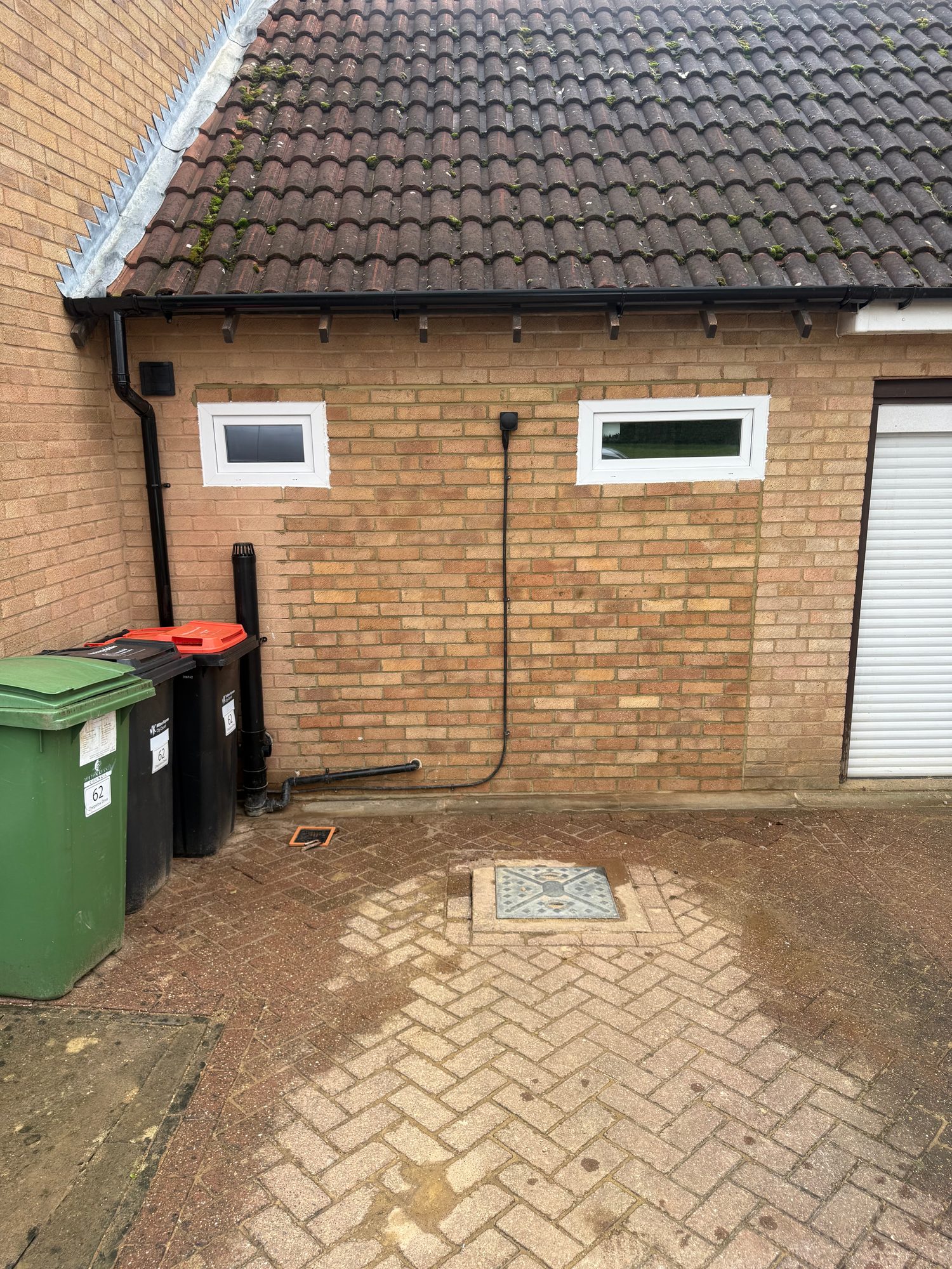 External brick wall with two white-framed windows, black rainwater goods and a tiled roof, with brick paving and drainage covers at ground level
