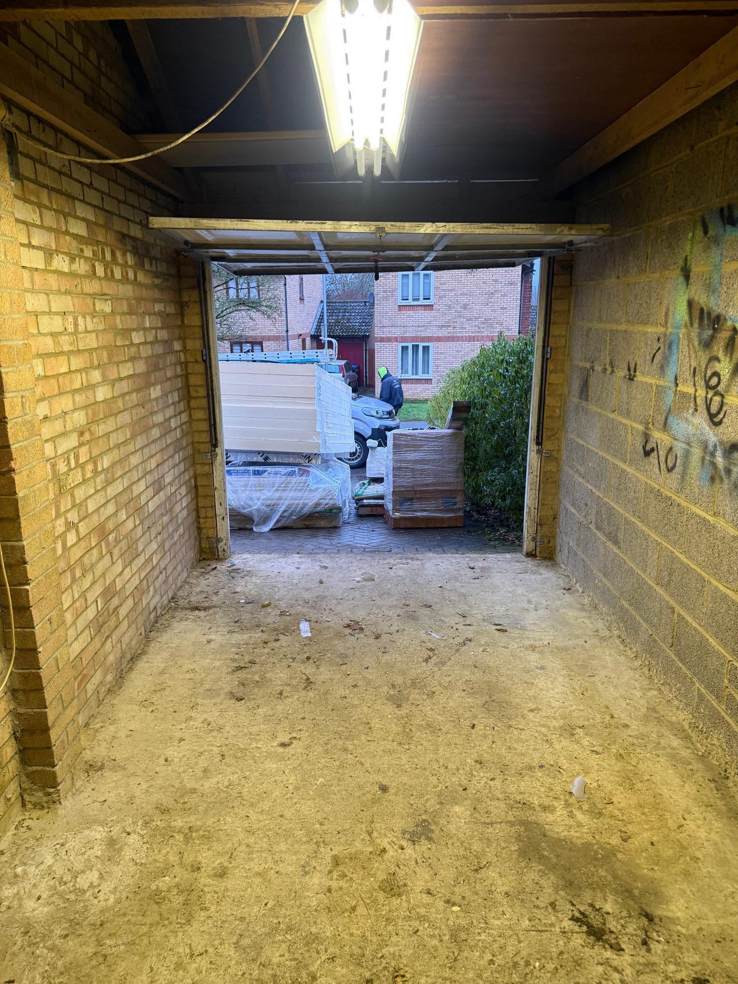 Interior view of a brick garage with the garage door open, showing a concrete floor, overhead lighting and wrapped building materials positioned outside