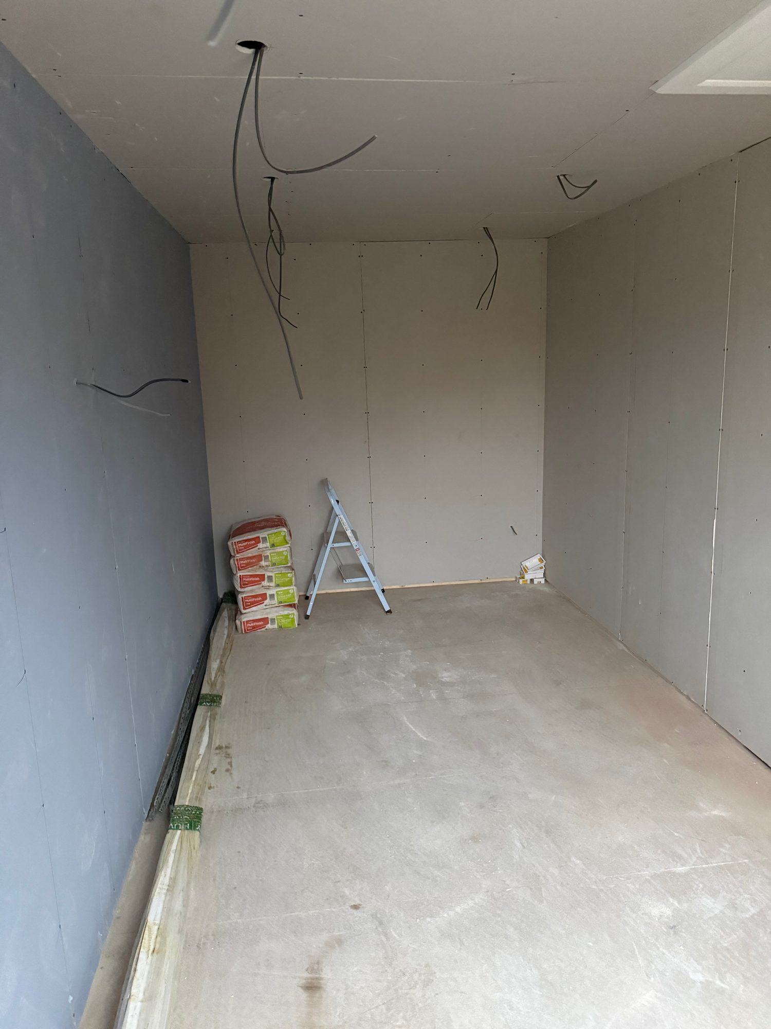 Partially finished room with plasterboard walls and ceiling, exposed electrical cables, a step ladder and building materials on a concrete floor