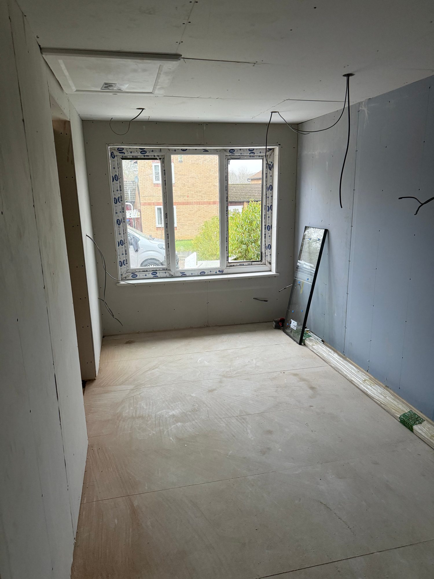 Interior room with plasterboard-lined walls and ceiling, a fitted three-section window, exposed electrical cables and a plywood-covered floor