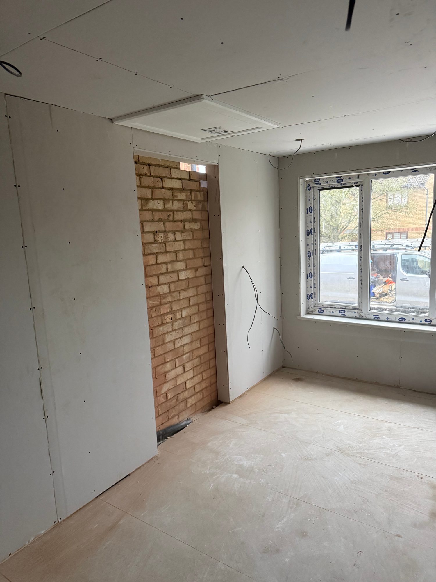 Interior room with plasterboard-lined walls and ceiling, a fitted window, exposed electrical cables, an open doorway and a plywood-covered floor