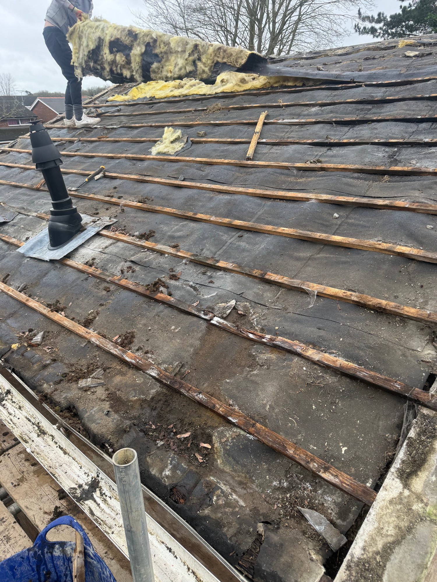 Pitched roof with tiles removed, exposing the underlay and timber battens, with sections of yellow insulation material visible and a roof vent remaining in place