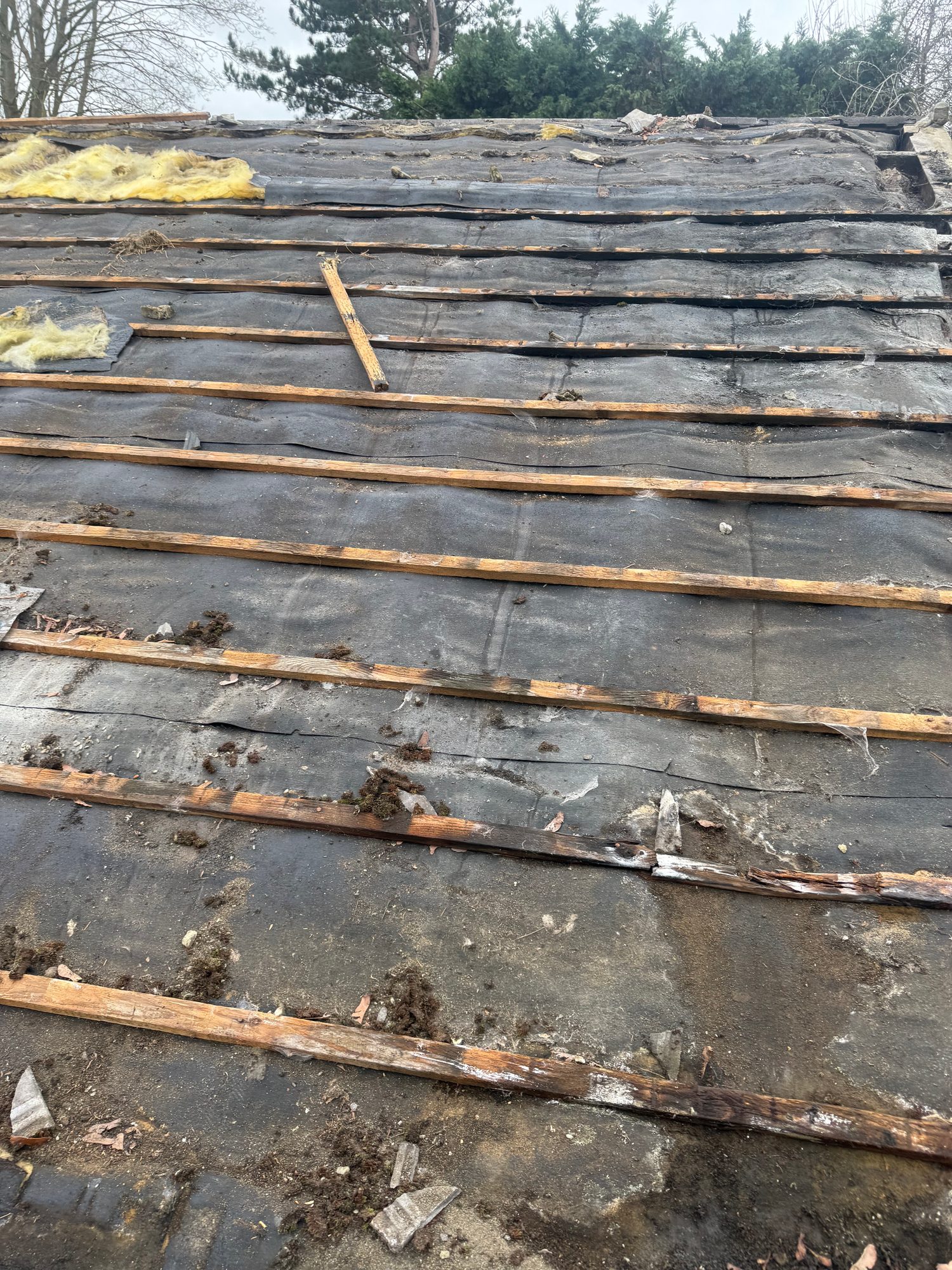 Close view of a pitched roof with tiles removed, showing the exposed black underlay and rows of timber battens, with loose debris and sections of yellow insulation visible
