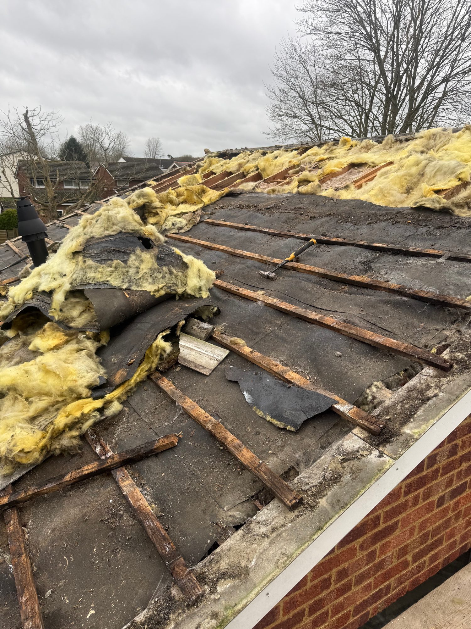 Pitched roof with tiles removed, showing exposed timber battens, black underlay and extensive yellow insulation material, with loose roofing debris and a roof vent visible