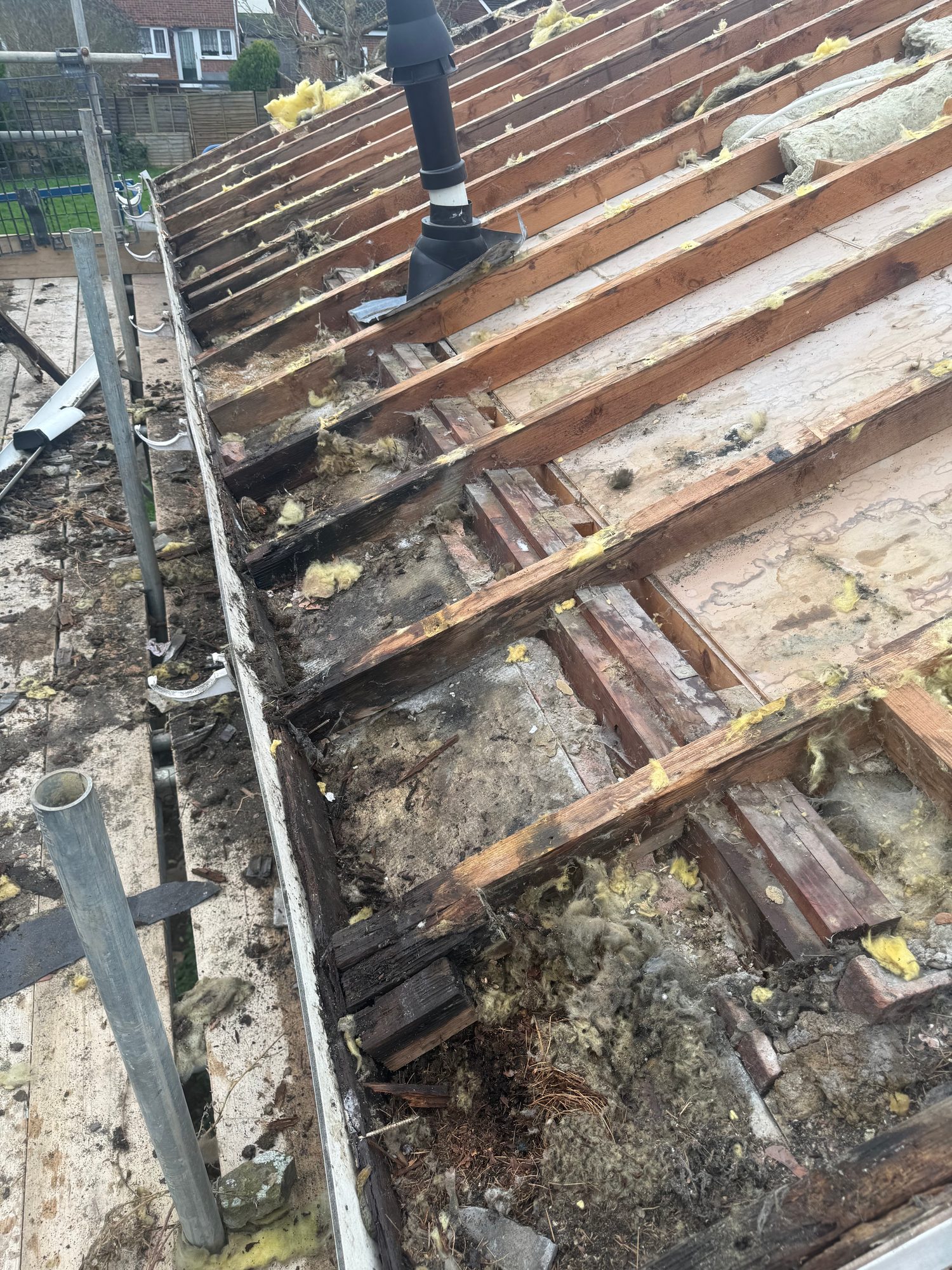Pitched roof with tiles removed, exposing timber rafters, underlay and remaining insulation, with a roof vent and scaffolding visible