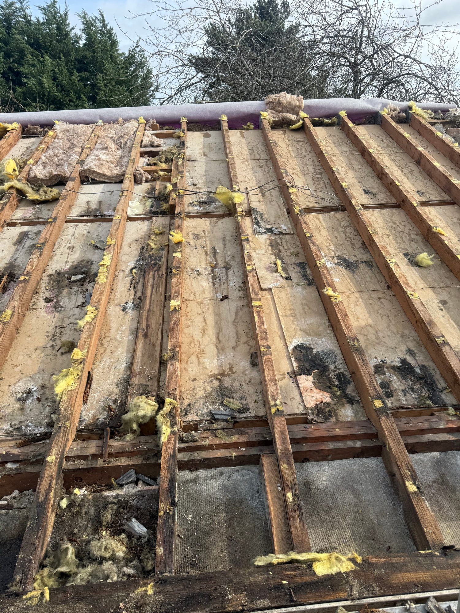 Pitched roof with tiles removed, showing exposed timber rafters, roof boards, remaining insulation and sections of roofing underlay