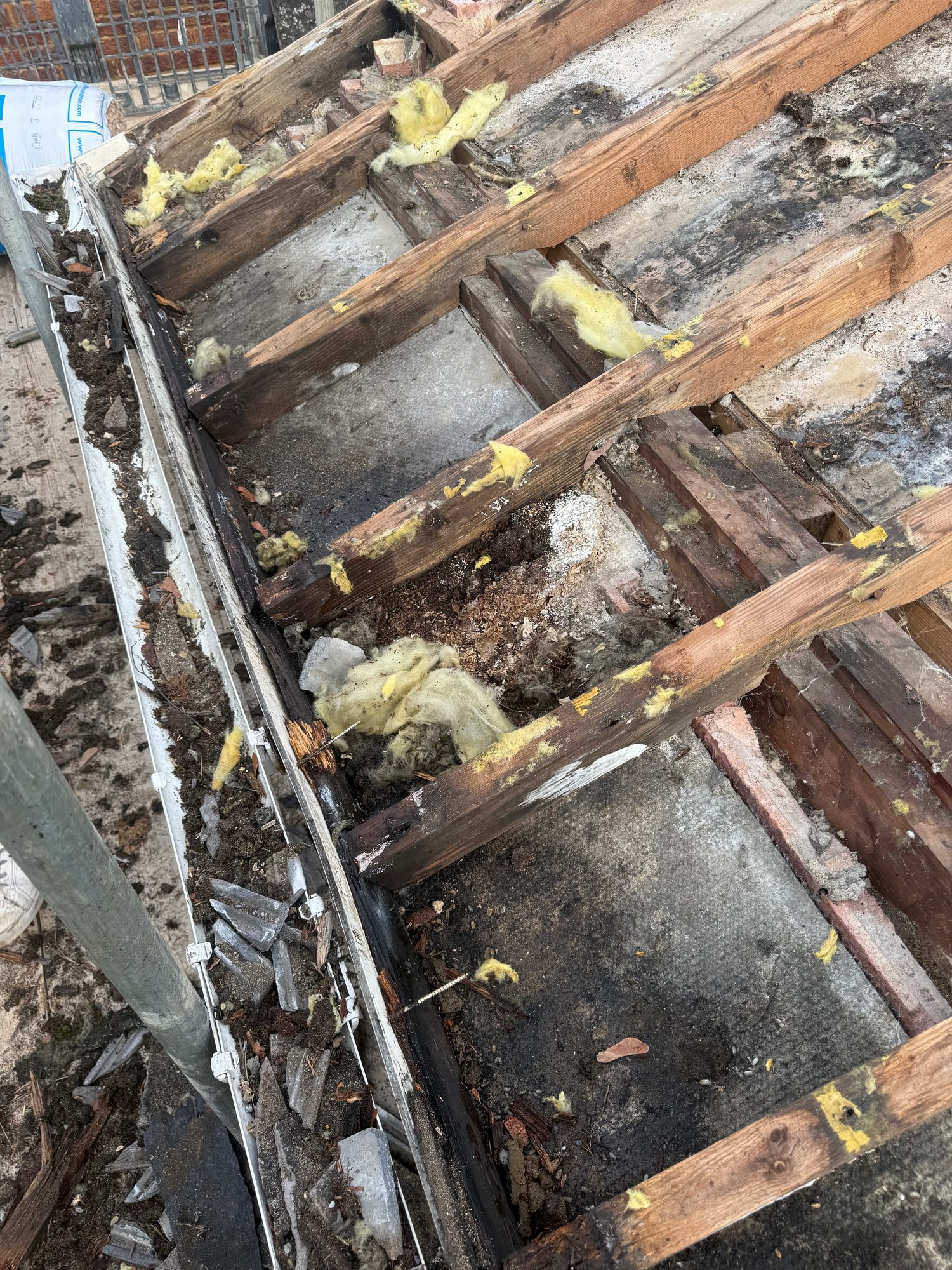 Close view of a stripped roof showing exposed timber rafters, remaining yellow insulation, roof covering material and accumulated debris along the roof edge