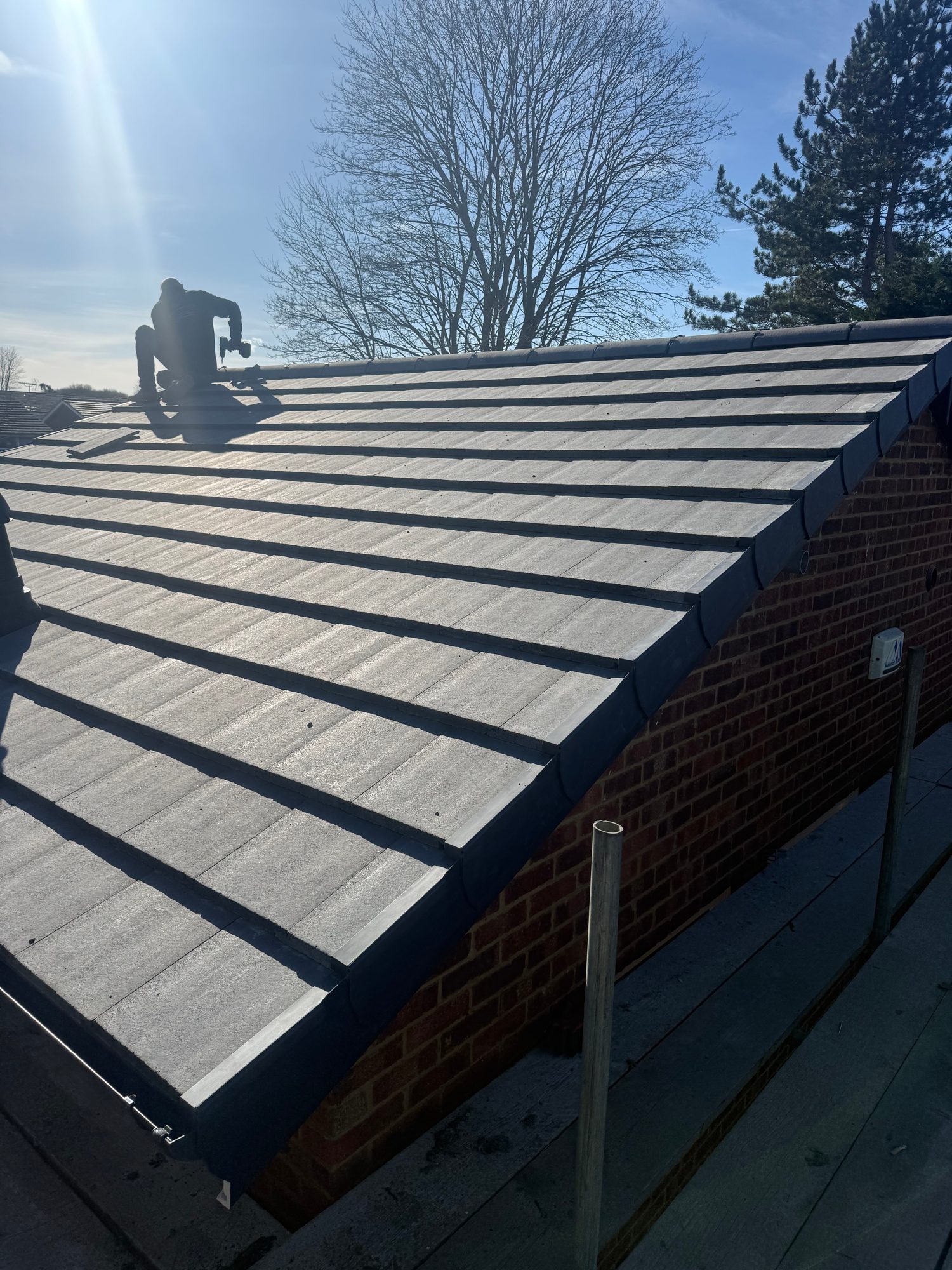 Pitched roof with grey tiles installed across the roof slope, finished with ridge tiles and dark verge detailing, with a worker visible near the ridge and scaffolding around the building