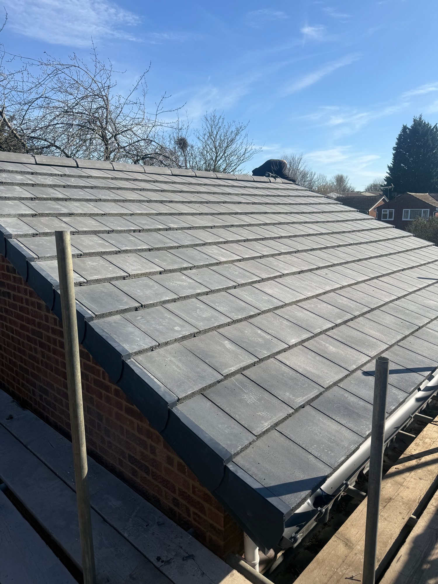 Wide view of a completed pitched roof covered with evenly laid grey tiles, with dark perimeter detailing, guttering and scaffold visible around the roof