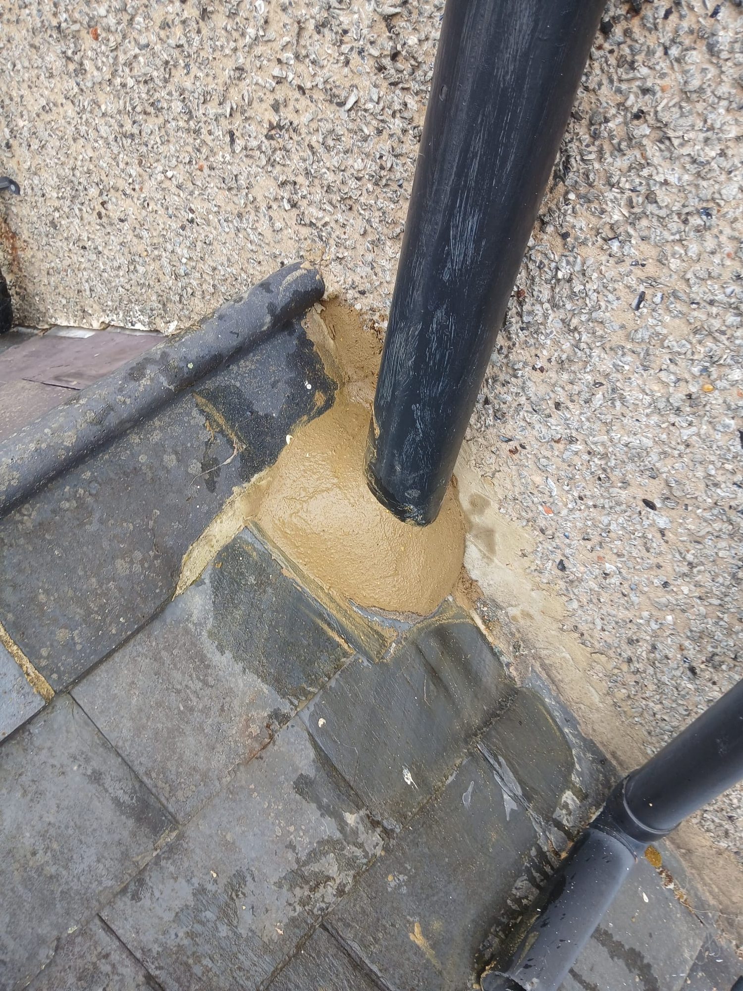 Black roof pipe passing through dark tiles with mortar applied around the junction between the pipe, roof and textured wall
