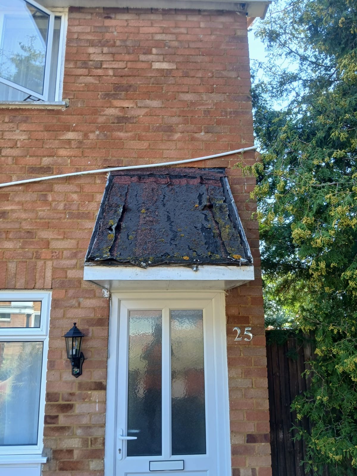 Brick house entrance with a small projecting roof canopy above a white glazed front door, showing a dark weathered roof covering with patches of moss