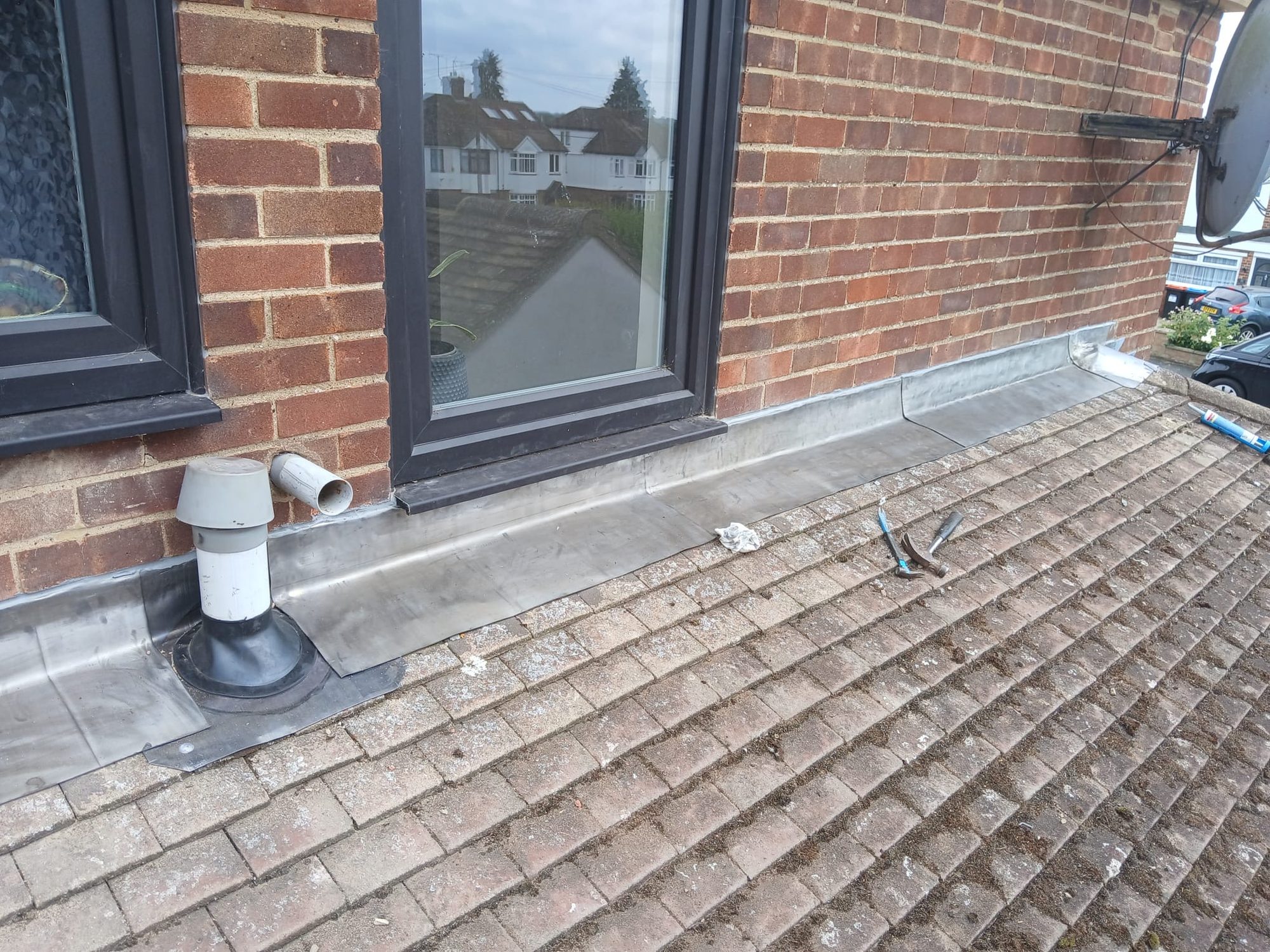 Lead flashing installed around two windows, a ventilation pipe and the base of a red brick wall above a tiled roof