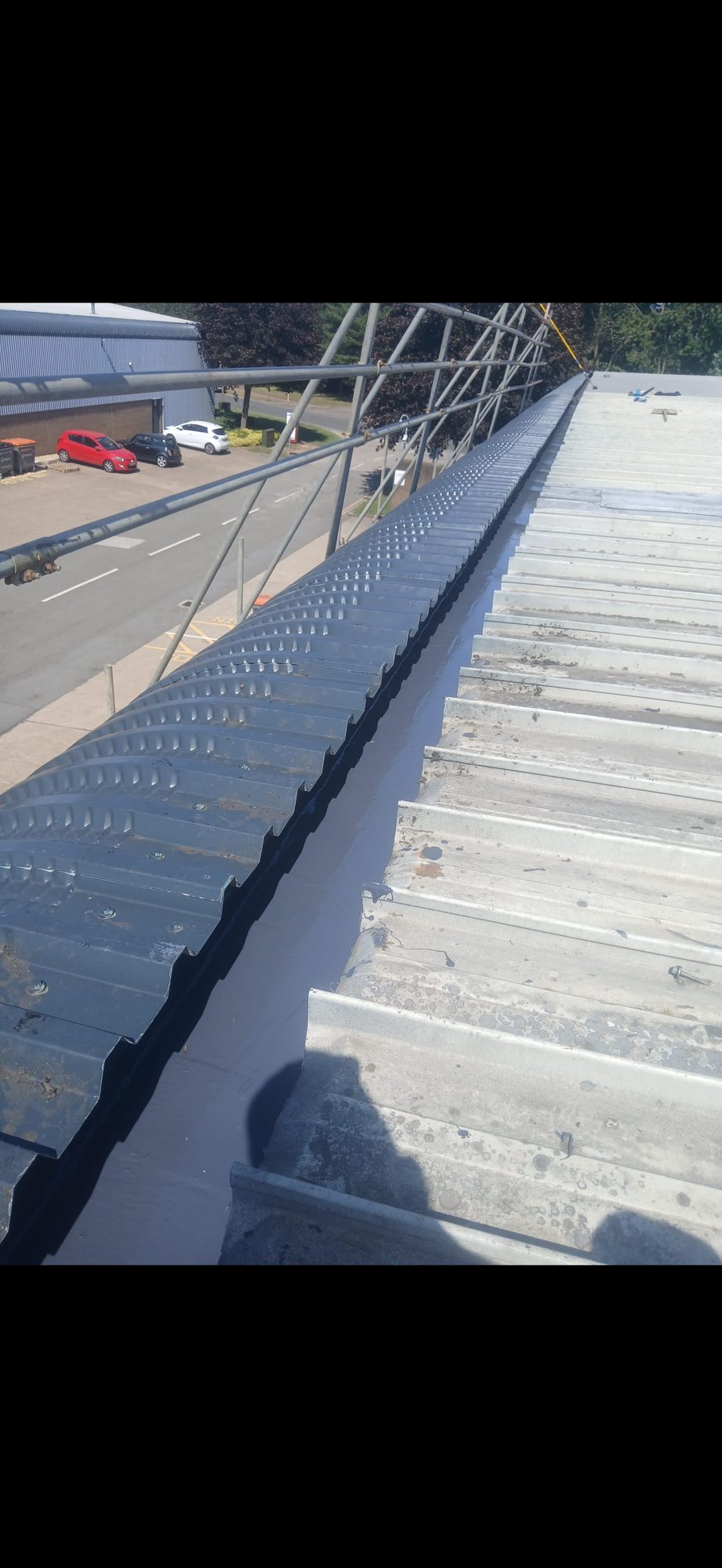 Elevated view along a black-coated roof channel between dark corrugated panels and a light metal roof beside scaffolding