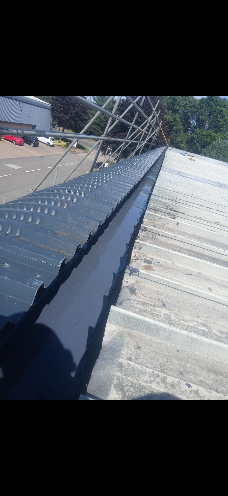 Long black waterproofed roof junction running between dark and light corrugated roofing with scaffolding at the side