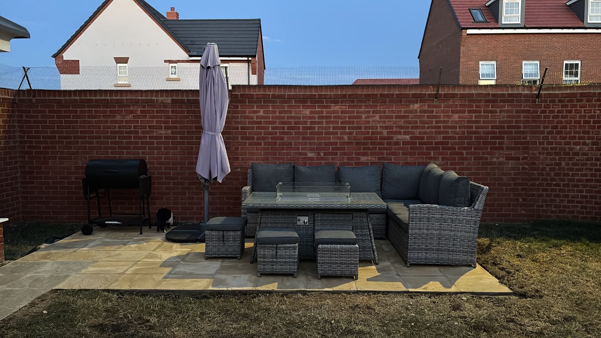Wide view of a paved seating area with grey outdoor furniture, a glass-topped table, parasol and barbecue