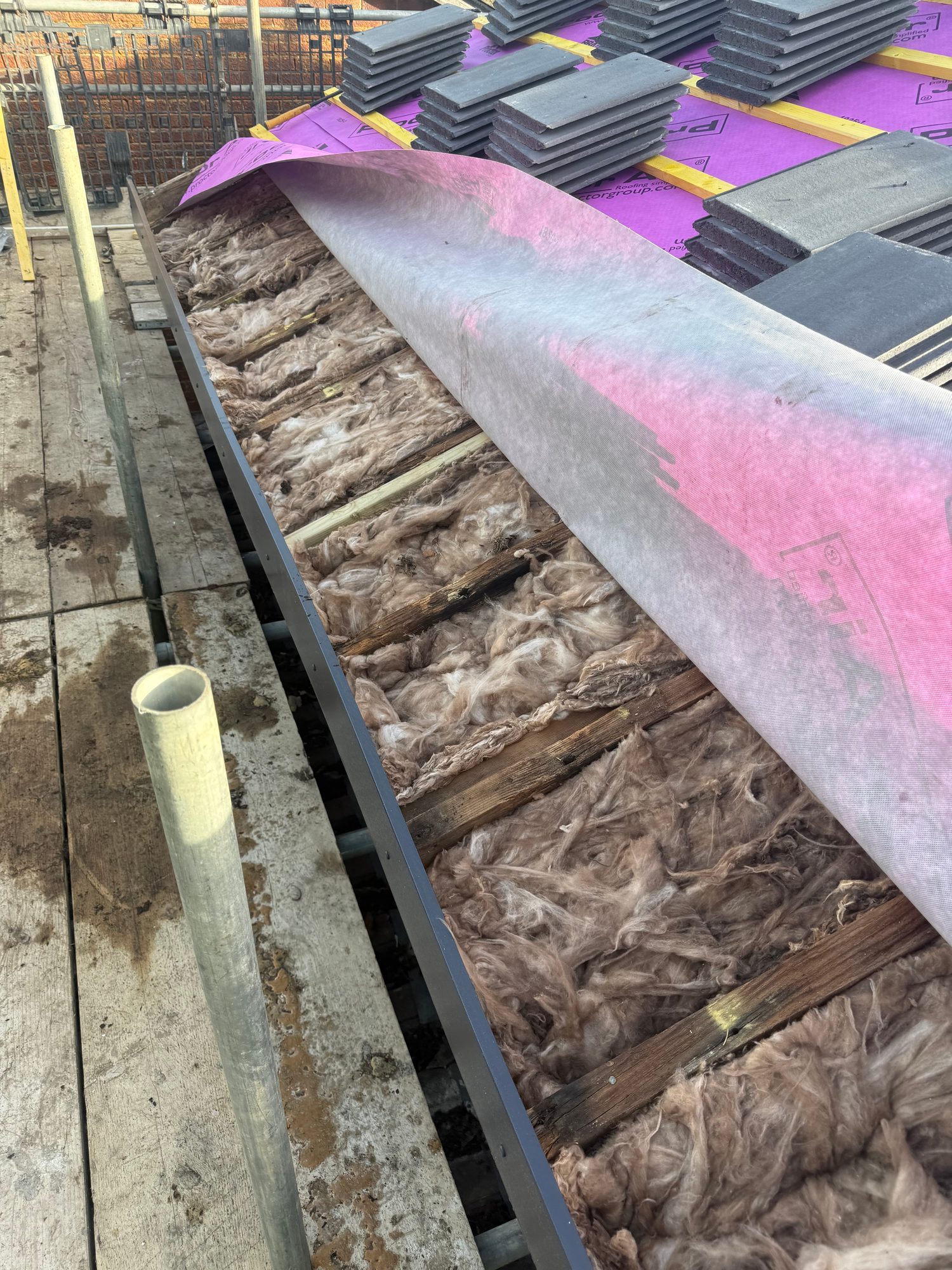 Roof section showing a purple roofing membrane over timber battens, exposed mineral wool insulation beneath the membrane and stacks of dark roof tiles ready for installation