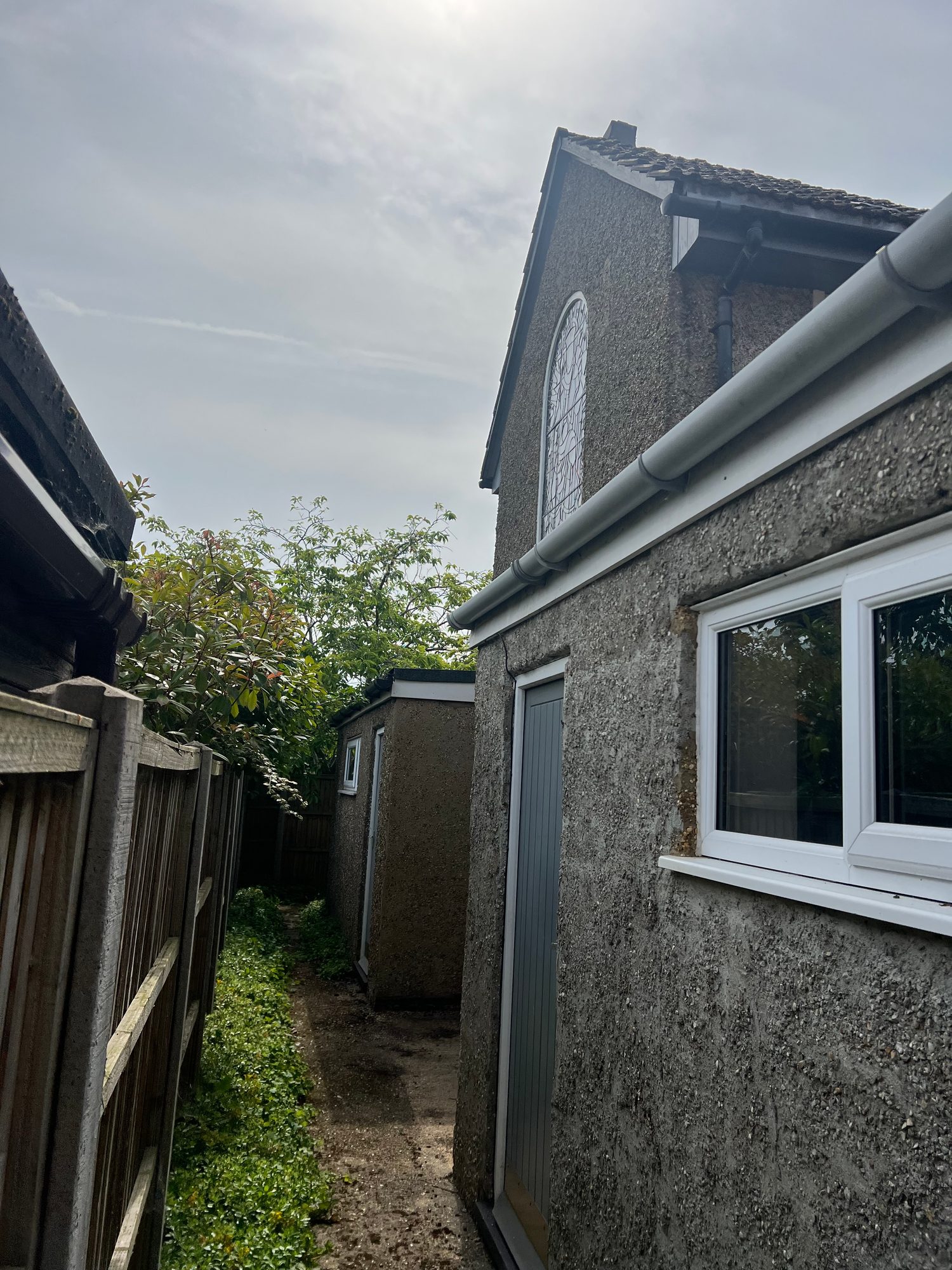 Narrow side passage beside the rendered church building showing a roof gutter, window, door, timber fencing and planted vegetation