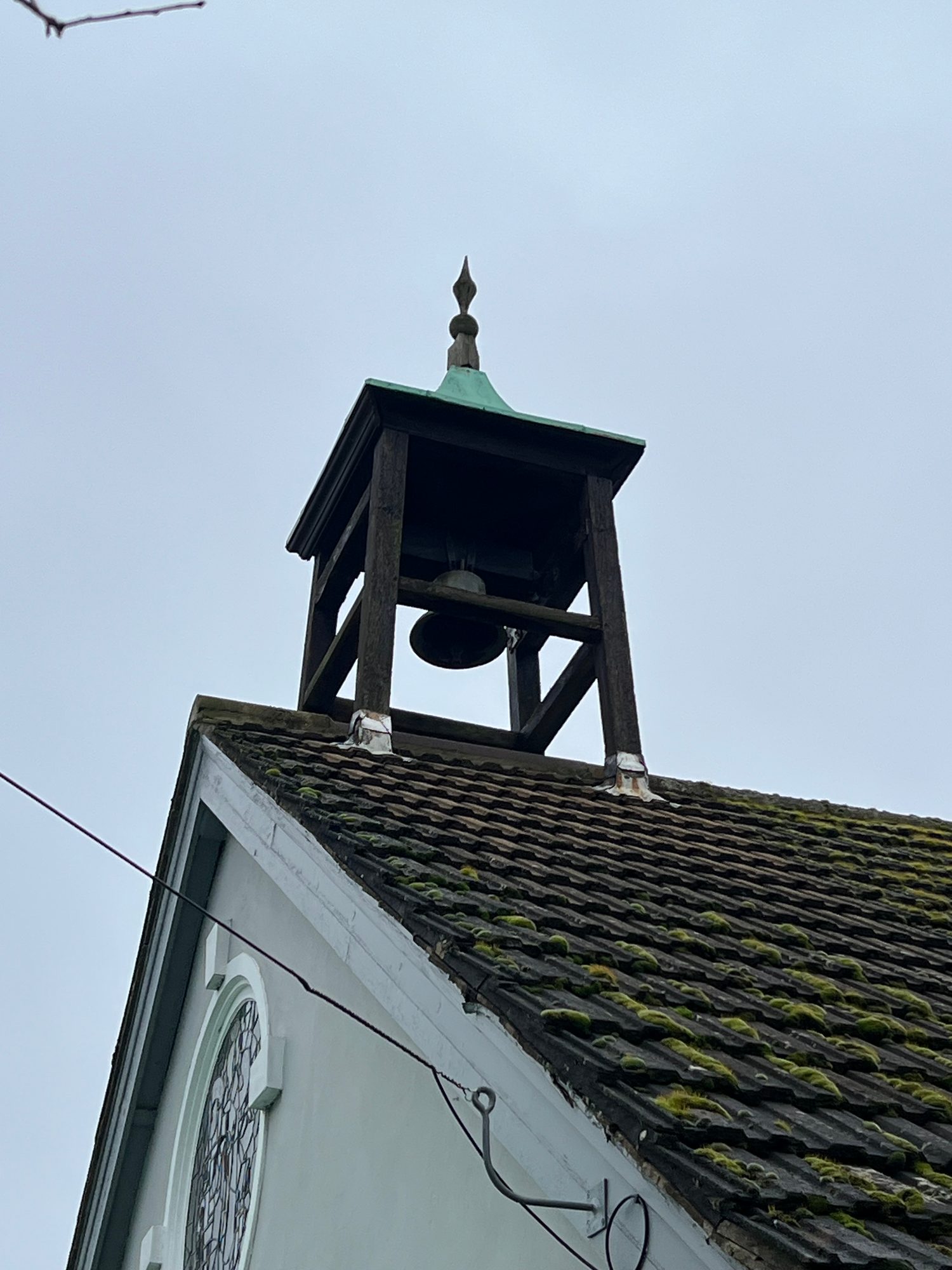 Close view of a timber bell tower above a pitched roof with extensive moss growth, with a circular window visible on the white gable end