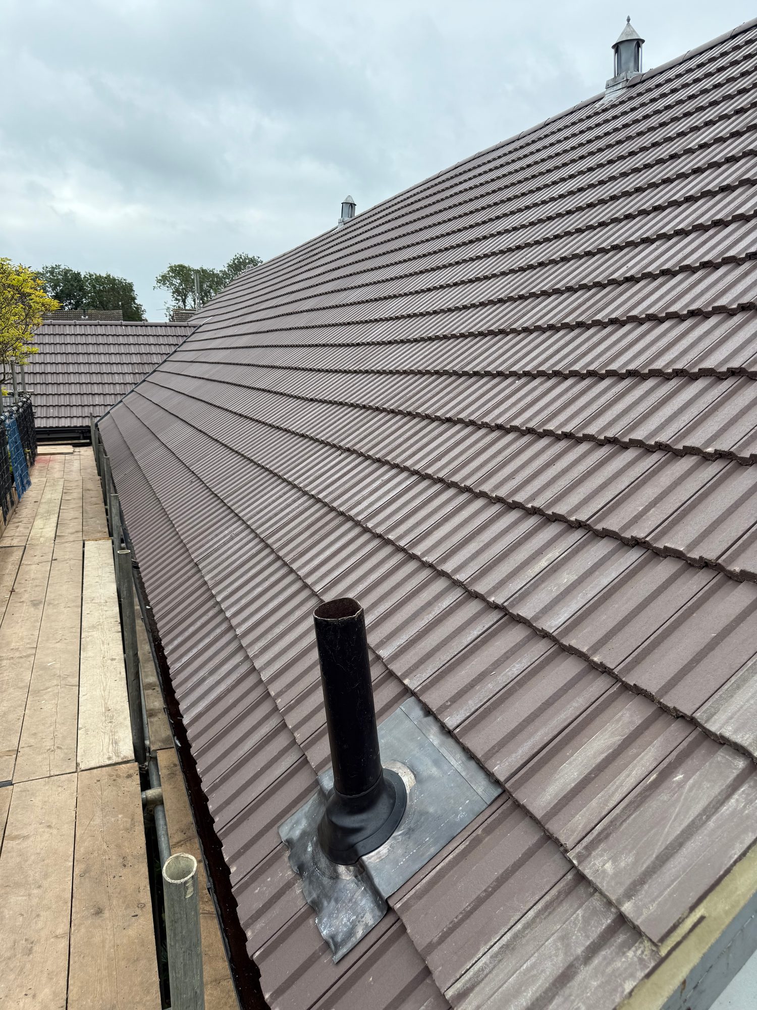 Completed tiled roof with a black vent pipe projecting through the tiles, surrounded by visible metal flashing, with scaffolding and a timber walkway along the roof edge