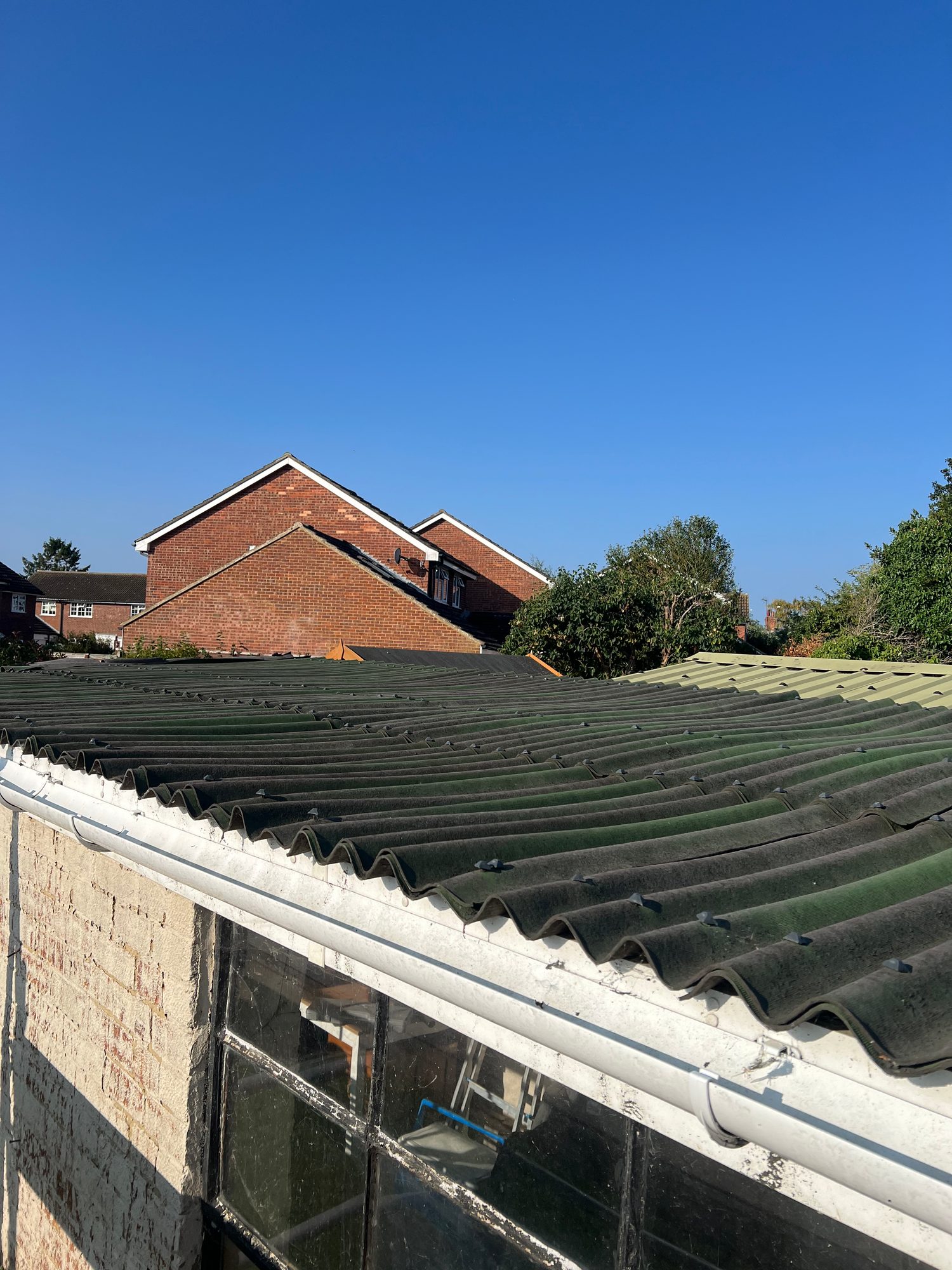 Existing corrugated roof covering with a white gutter along the roof edge, showing weathered roof panels and surrounding brick buildings