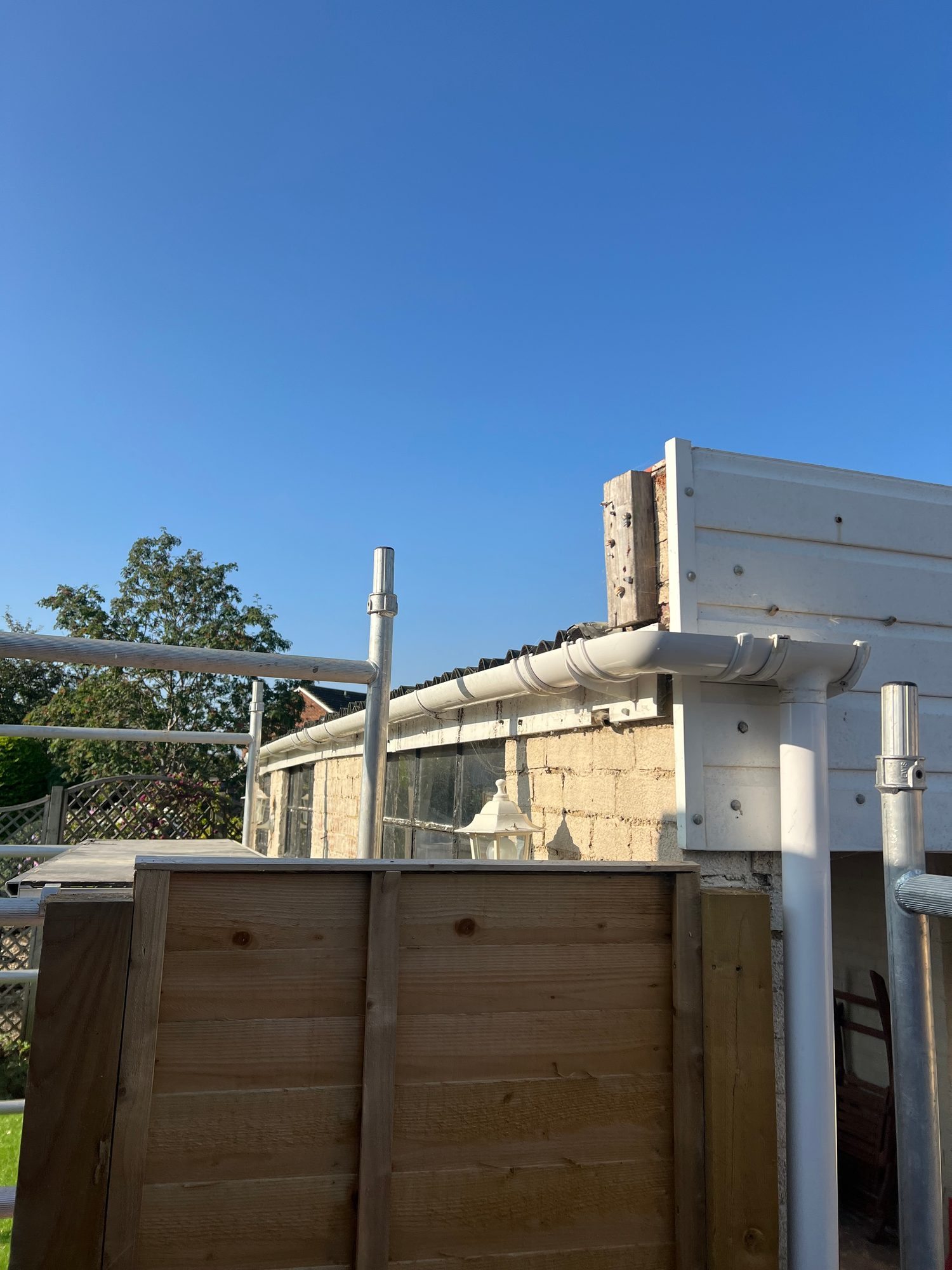 Existing roof edge with corrugated roof panels, white guttering and downpipe, with scaffolding positioned alongside the building