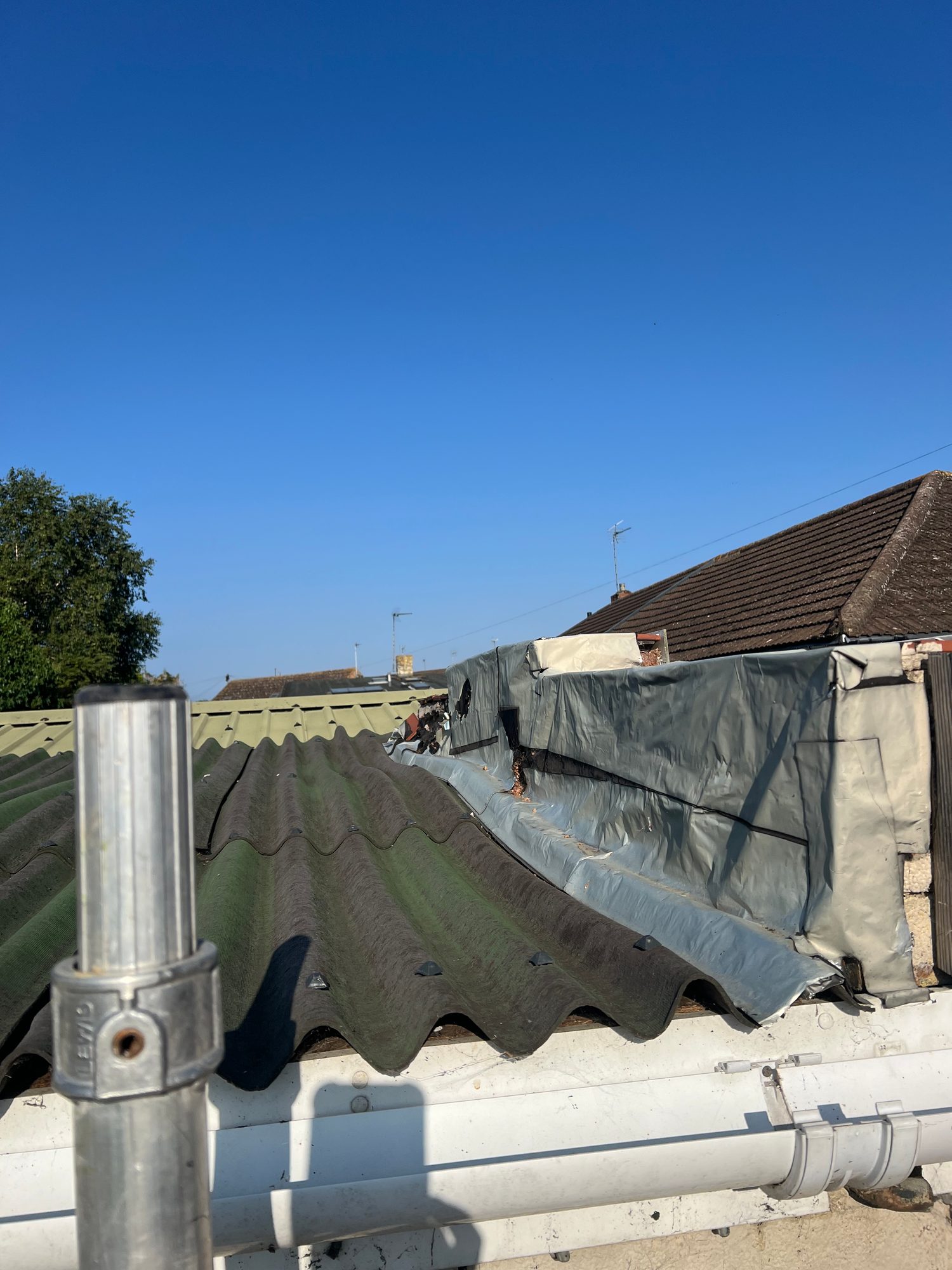 Existing roof area with corrugated roofing sheets, temporary-looking protective sheeting along the adjoining roof edge and visible guttering, photographed before works begin
