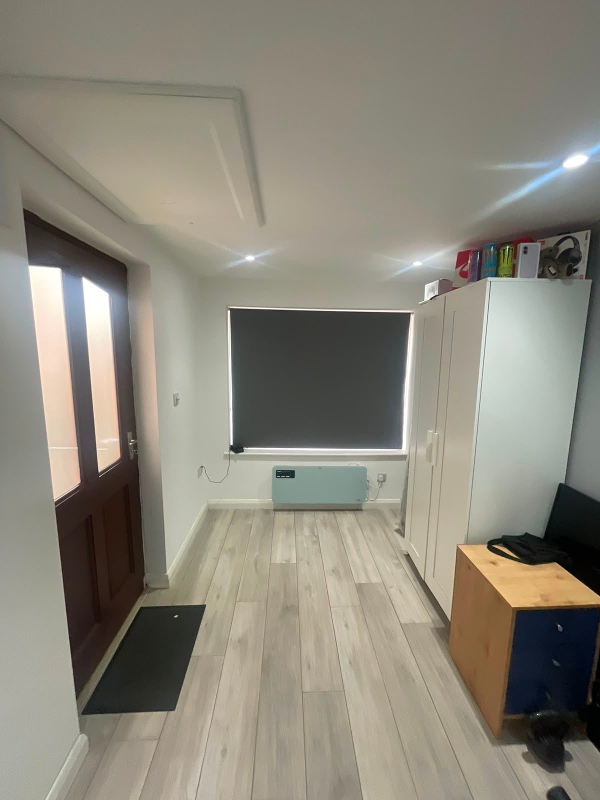 Refurbished room with light wood-effect flooring, white and grey walls, recessed ceiling lights, a window with a roller blind and fitted storage
