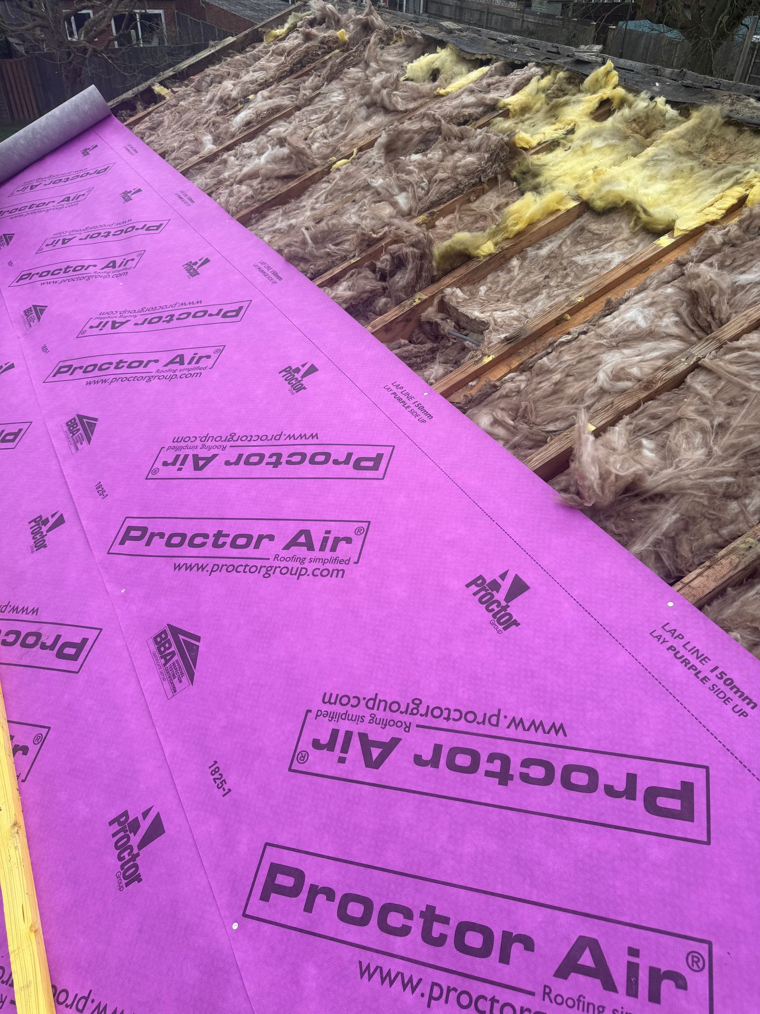 Pitched roof showing purple roofing underlay installed across part of the roof, with exposed mineral wool insulation and timber rafters visible on the remaining section