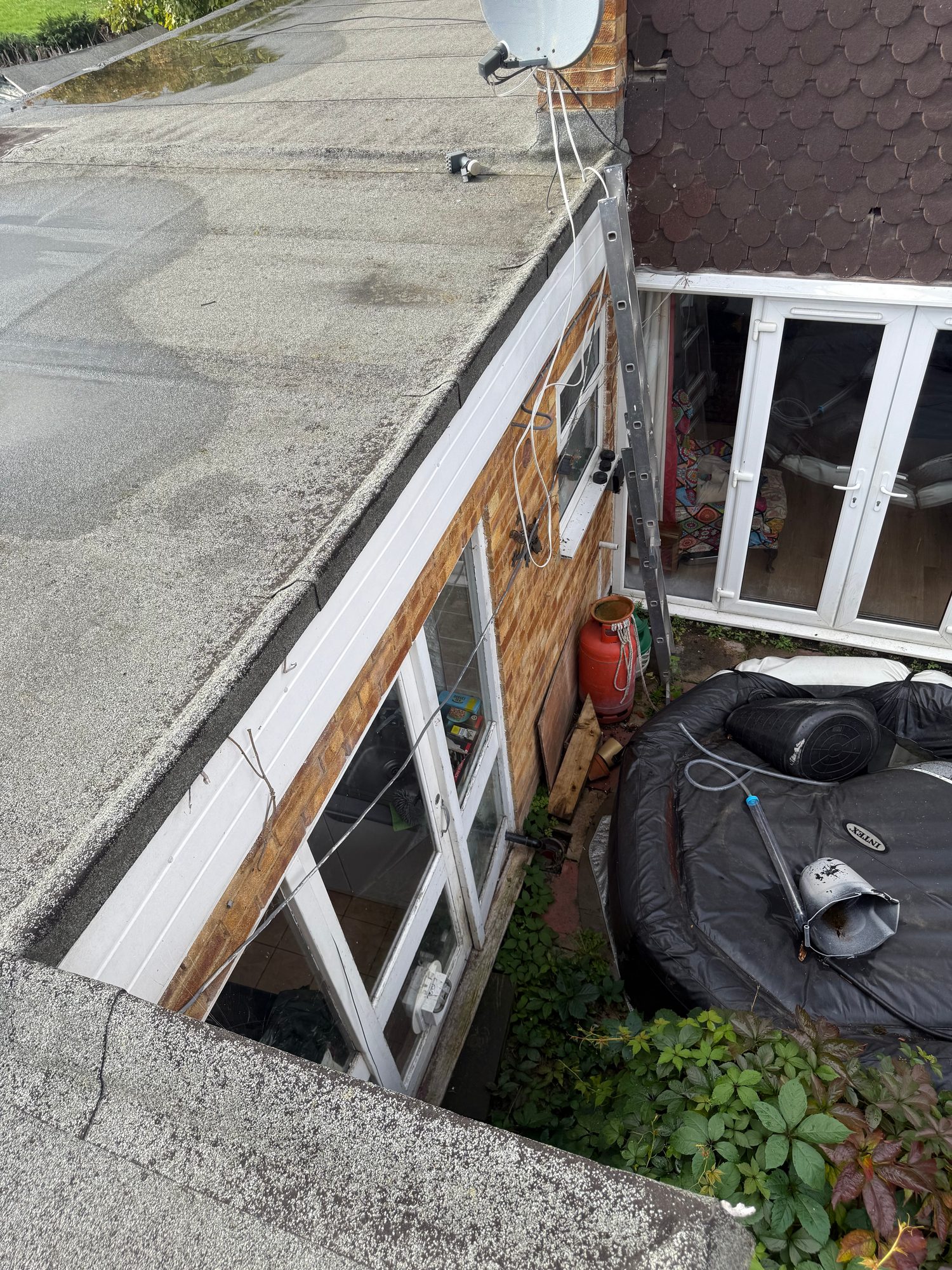 View of a flat roof with visible roof covering and seams, a white roof edge trim, an adjoining brick wall with windows and external cables, and a ladder positioned against the wall