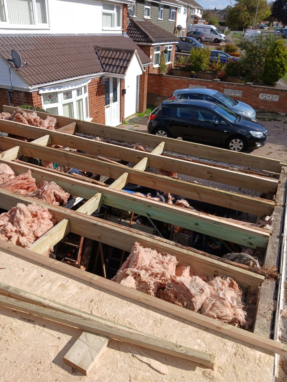 Exposed roof structure showing timber joists, cross supports and sections of pink insulation, with neighbouring houses and parked cars visible beyond