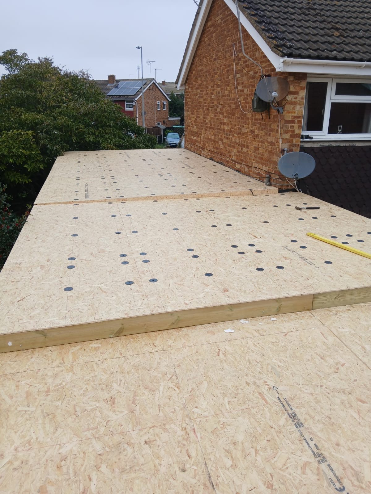 Newly installed OSB boards forming a roof deck beside the existing brick house, with fixings visible across the board surface and neighbouring properties in the background