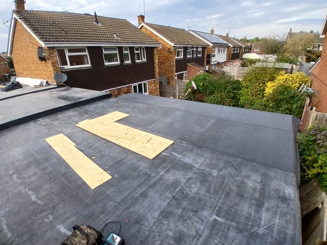 Wide view of a flat roof during installation, with timber boards positioned on the roof surface and neighbouring residential properties visible beyond