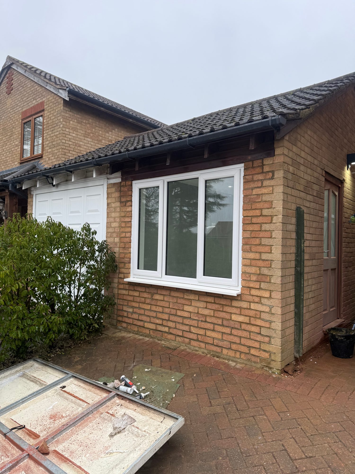 Exterior view of a brick building showing a newly fitted white-framed three-panel window, with the garage door and tiled roof visible
